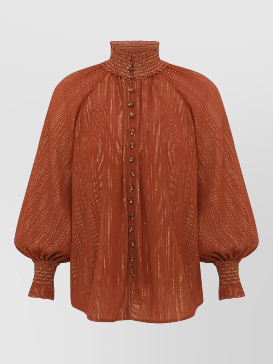 Zimmermann Gathered blouse — The rust metallic mini mirrors the jacket’s sculpted drama and warm brown palette, creating a rich, tonal anchor that respects the outerwear’s architectural volume without competing in length.