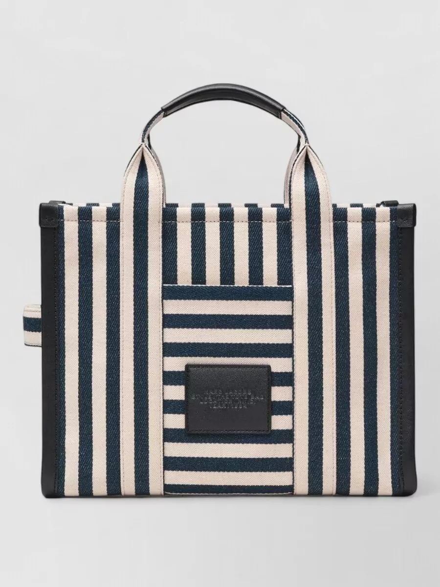 Marc Jacobs the tote bag medium canvas stripe tote bag — The structured navy-and-cream tote leans into the Riviera stripe story and scale of the trousers, giving a preppy, practical finish that still reads luxury off-duty.