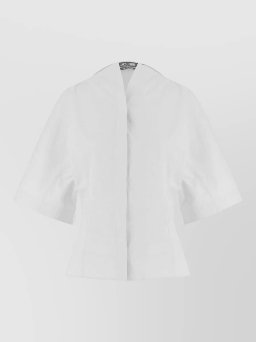 Jacquemus Cotton long-sleeve blouse — The Jacquemus structured short‑sleeve blouse keeps the ivory-on-ivory palette, matches the tailored quiet-luxury mood, and balances the long tailored shorts with a clean, sculpted summer silhouette.