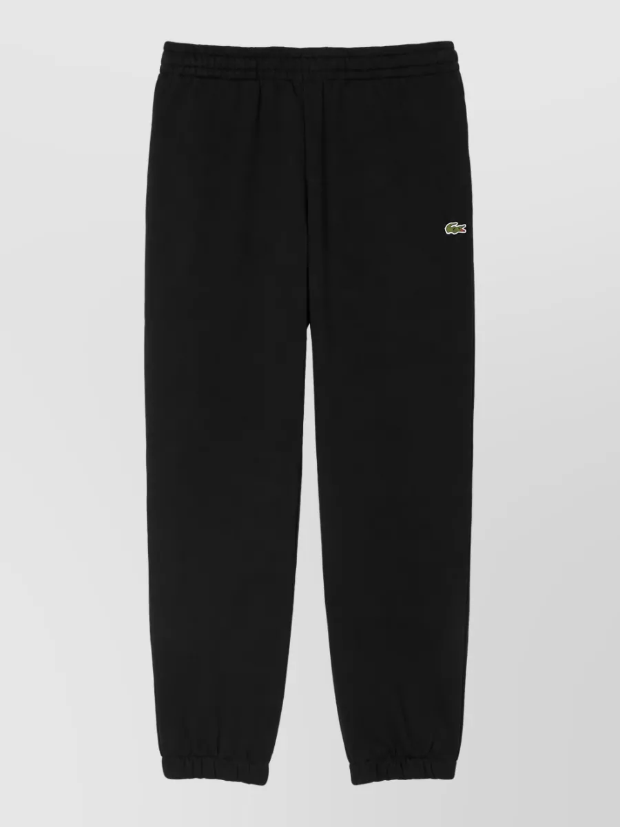 Lacoste Regular-fit track pants — The black Lacoste cuffed track pant keeps the palette tonal with the black mock-neck, matches the sport-preppy mood, and creates a clean, uninterrupted line for a streamlined athleisure set.