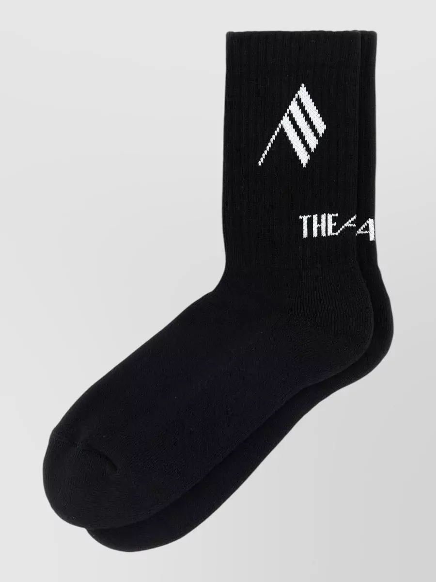 The Attico Cotton socks — The black Attico logo socks peek out just enough above the teal sneakers to tie in the black bag and jacket, giving a deliberate, styled finish without crowding the print.