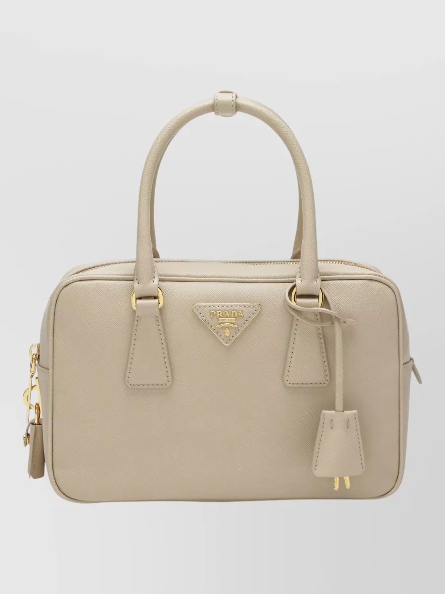Prada Saffiano leather top handle bag — G, the structured warm-beige Prada top-handle, mirrors the blazer’s refined lines and neutral palette while matching its price tier and formality for a coherent boardroom-ready finish.
