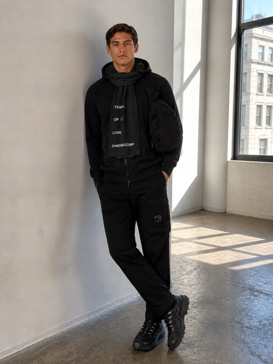 Head-to-toe C.P. Company in deep black and charcoal keeps this outfit tight and urban, with the goggle hoodie and tapered cargos doing the heavy lifting on detail. The monochrome Salomon runners and technical sling bag keep everything practical and fast, while the gauzy Rick Owens scarf adds just enough edge if you want a graphic accent. It is a clean, city-ready uniform that feels considered without trying hard.