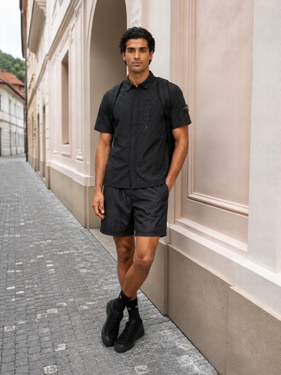 Head-to-toe Stone Island nylon in deep charcoal and black creates a tight, technical story that feels urban and low-key. The crinkled shirt and deck shorts read like a modern two-piece set, with the black sneakers and socks keeping the proportions clean and athletic. It is a sharp, easy uniform for warm city days that can move from coffee runs to late terrace drinks without changing a thing.