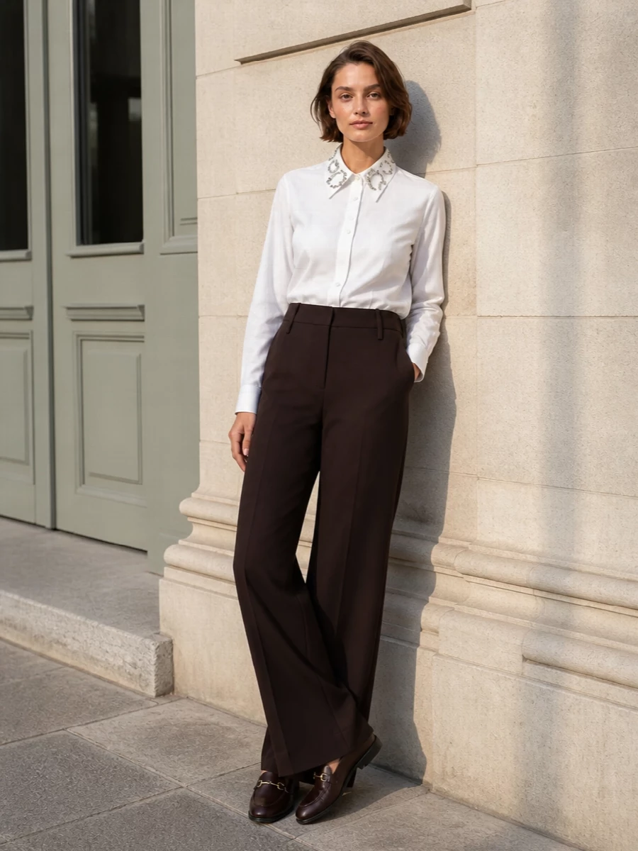 A crisp white jeweled-collar shirt tucked into high-waisted espresso wide-leg trousers creates a long, precise line that feels considered from morning meetings through dinner. The chocolate loafers ground the palette in rich neutrals, while the structured camel-toned shoulder bag adds a warm counterpoint and quietly echoes the gold hardware. This route leans into tailored separates rather than a dress, which better suits the preppy, boardroom-to-city character of the horsebit loafer.