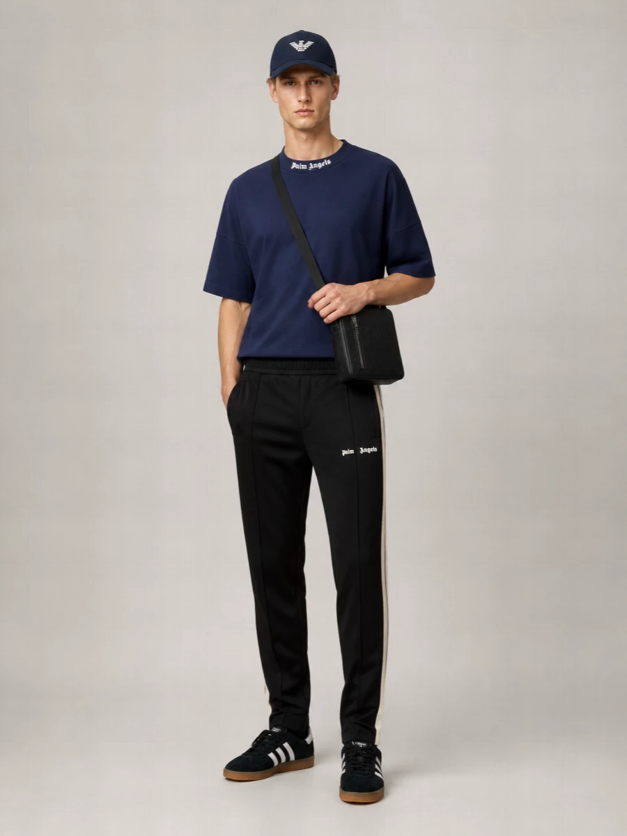 A navy Palm Angels tee and black striped track pants create a clean, athletic base that feels modern and relaxed under the Emporio Armani cap. Black and white terrace sneakers keep the lines streamlined while the Gucci crossbody brings a quiet hit of luxury to the street-sport mix. The palette stays tight in navy, black and white, so the graphic branding reads sharp rather than loud.
