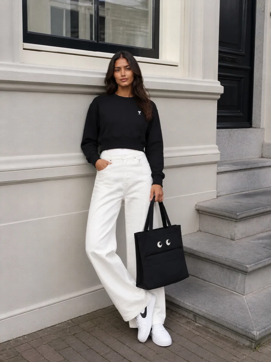 A cropped black Ami sweatshirt sits against structured white wide-leg denim for a crisp, collegiate take on streetwear. Monochrome Nike sneakers keep the lines clean and sporty, while the graphic Anya Hindmarch tote adds a witty preppy twist without sacrificing polish.