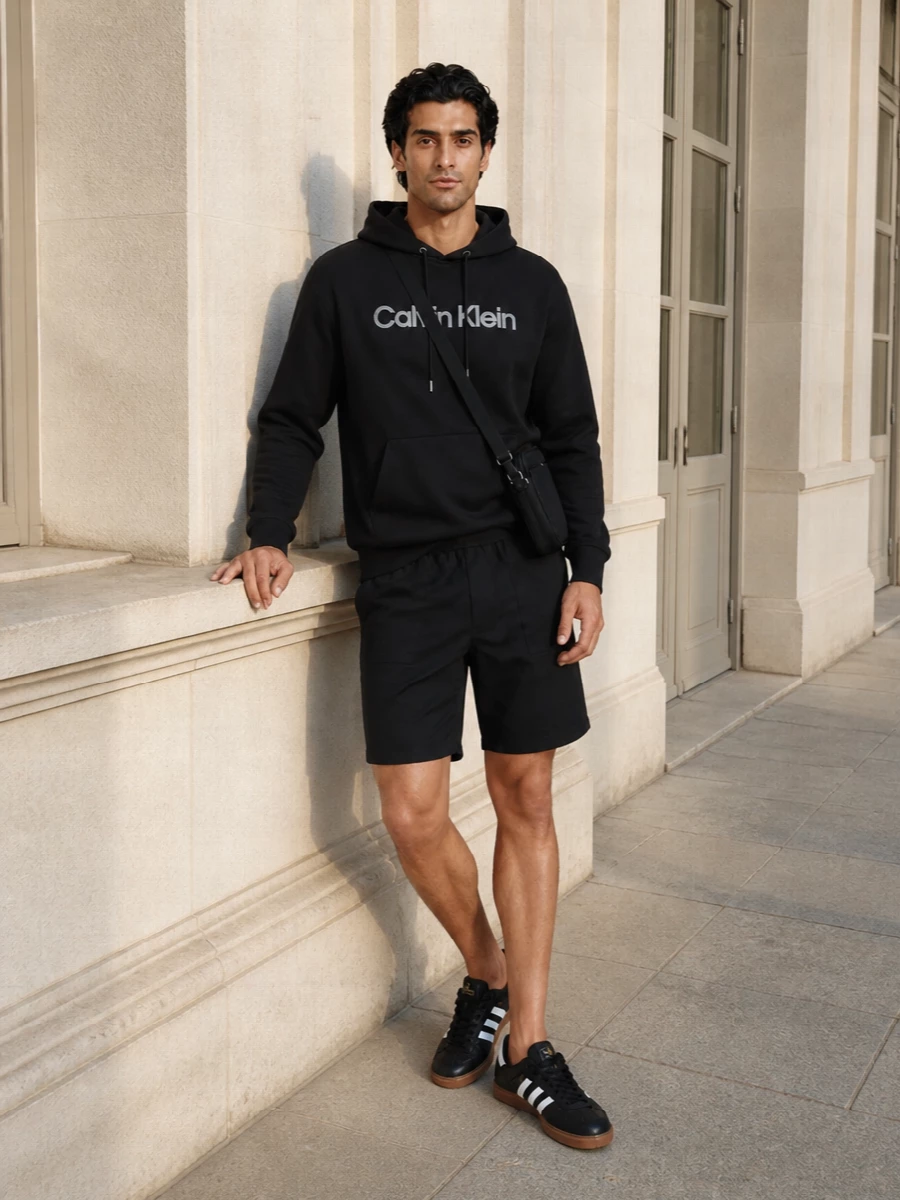A black Calvin Klein logo hoodie sits over matching clean-cut black shorts for a tight, minimalist base. Classic black-and-white Adidas sneakers keep the palette graphic but easy, while a slim black crossbody and simple belt add just enough structure for city days or travel. The result is a relaxed sport ensemble that feels intentional without trying too hard.