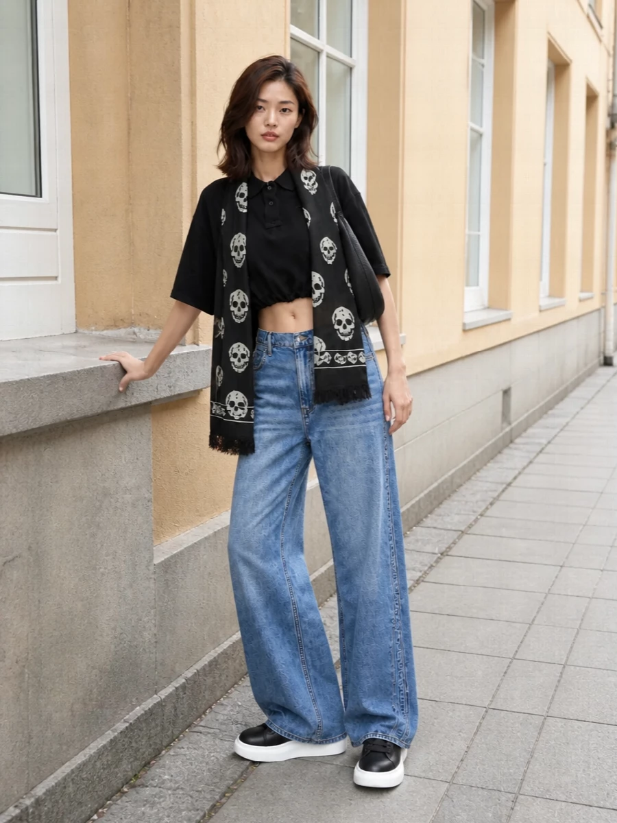 A cropped black polo sits over mid-wash wide-leg denim for a clean, slouchy streetwear take with a subtle Y2K lean. Chunky black and white sneakers bring height and attitude, while a sculptural black shoulder bag and a monochrome skull scarf sharpen the whole thing into a hard-working city uniform that moves easily from day to late night.