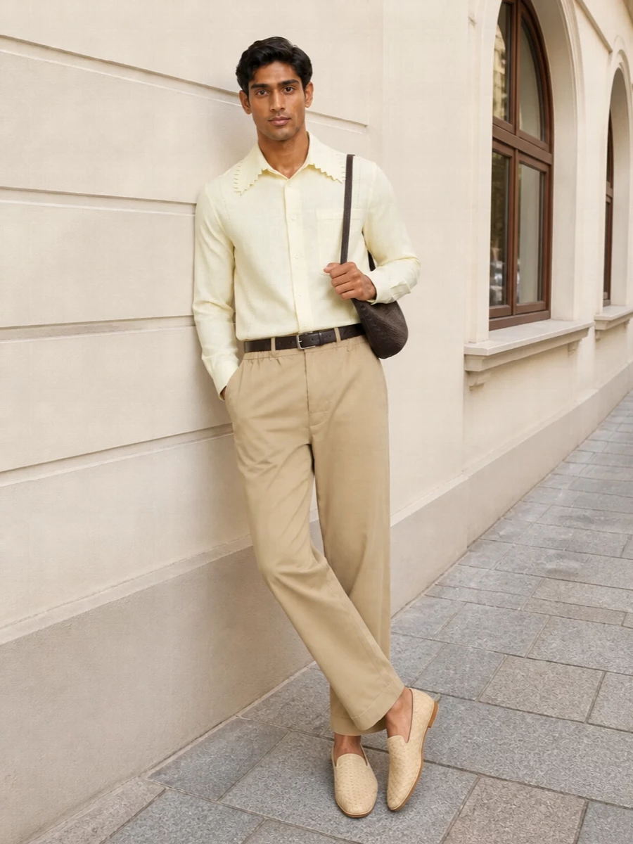 Sand trousers and a pale butter shirt create a soft, tonal column that feels relaxed but considered for warm city days. Woven slipper loafers keep the line clean and light underfoot, while dark espresso leather in the bag and belt adds just enough definition without breaking the quiet palette.