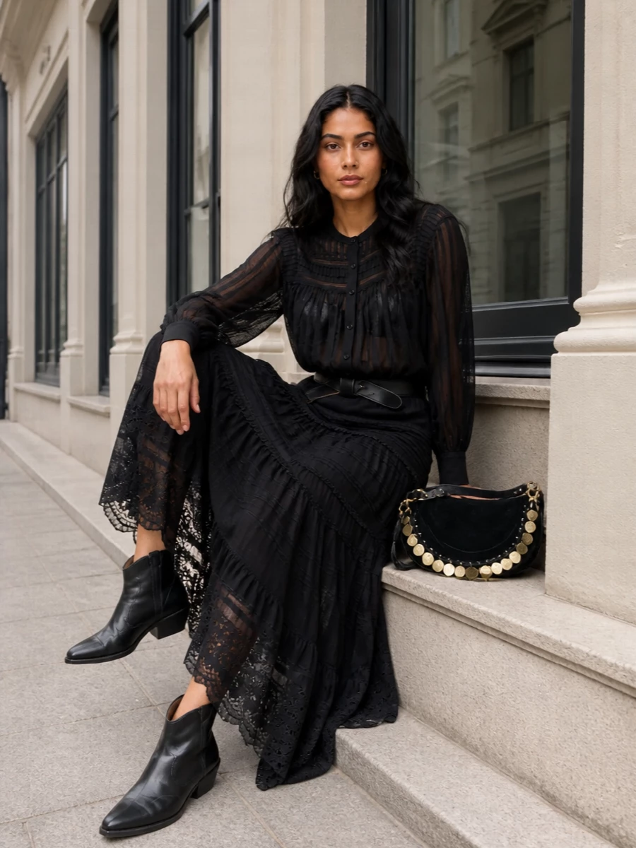 A sheer black peasant blouse pairs with the tiered black maxi to create a long, romantic silhouette that moves softly around the body. Western-inflected black boots and a coin-trimmed hobo bag sharpen the mood without disturbing the boho ease. A knotted leather belt quietly pulls the waist in, balancing all that volume with a considered, feminine line.