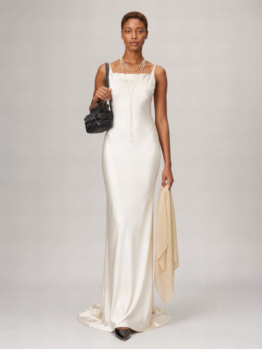 An ivory satin column, sharpened with black patent accessories, feels quietly commanding for evening without a trace of fuss. The pearl lariat traces the neckline and amplifies the gown’s vertical line, while the compact frame bag keeps the focus on that liquid, floor-grazing silhouette. It is modern, formal and intentionally spare, letting cut, texture and light do all the work.