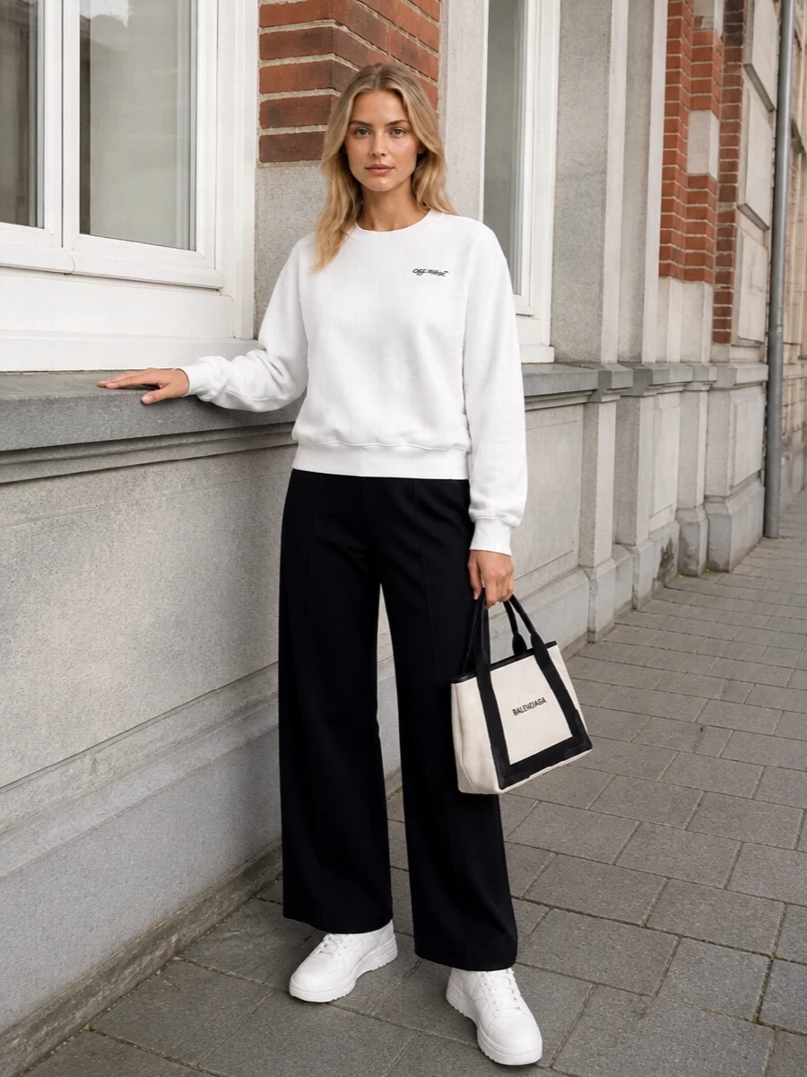 A soft-white logo sweatshirt meets high-rise black wide-leg track trousers for a monochrome take on sport that feels clean and intentionally slouchy. White court sneakers and an off-white structured mini tote keep everything crisp and graphic, so the ease reads styled rather than lazy. Ribbed socks peeking above the sneakers add just enough athletic detail to finish the set.