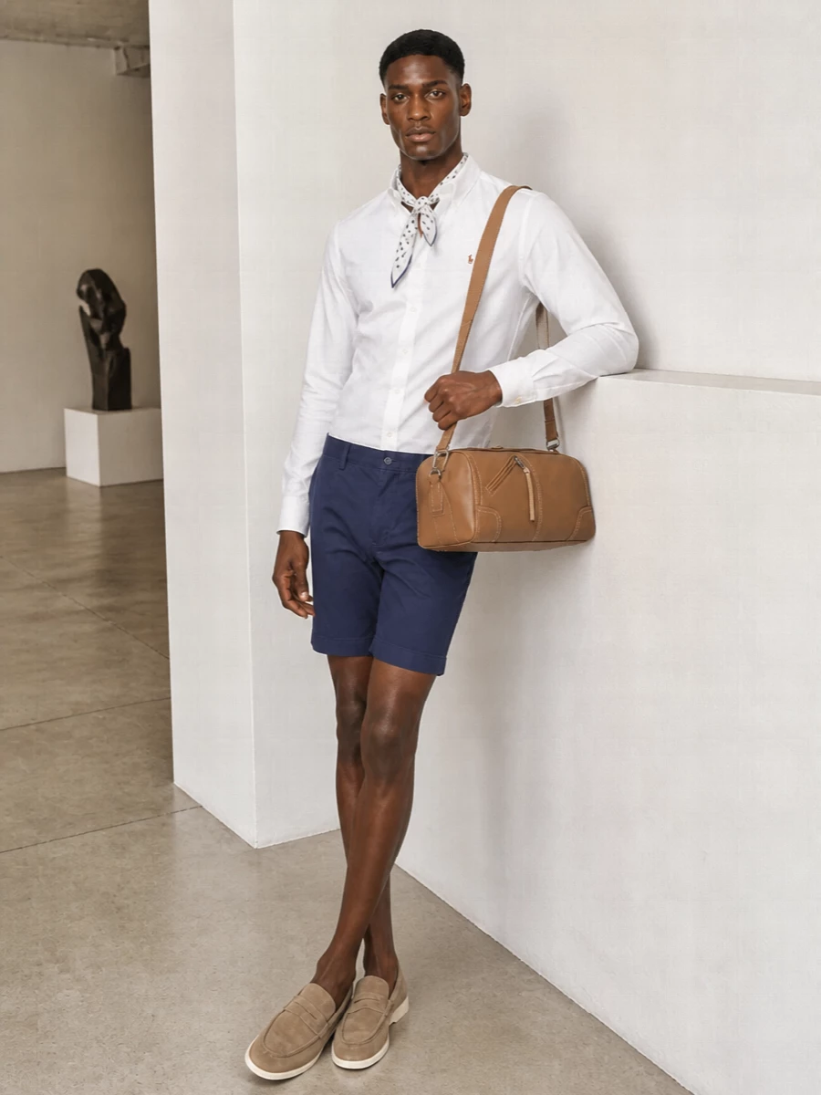 A crisp white Oxford meets navy chino shorts for that clean campus energy that still feels right at a terrace lunch. Taupe suede loafers keep it relaxed but refined, while the tan crossbody adds a useful touch of warmth. Finish with a neatly knotted dotted square for a small, intentional hit of pattern that stays firmly in the preppy lane.