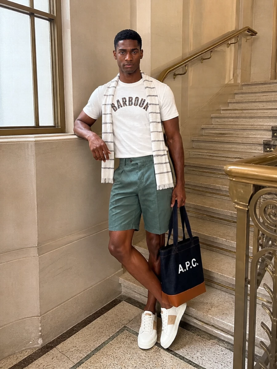 An off-white Barbour tee pairs with sage pleated Bermudas for a clean, collegiate take on warm-weather dressing. Off-white and camel-accented sneakers keep the lines sporty but refined, while the indigo A.P.C. tote and a fine striped scarf pull the palette into a relaxed, coastal-preppy mood. The proportions stay easy and regular, so the whole outfit feels considered without ever looking try-hard.