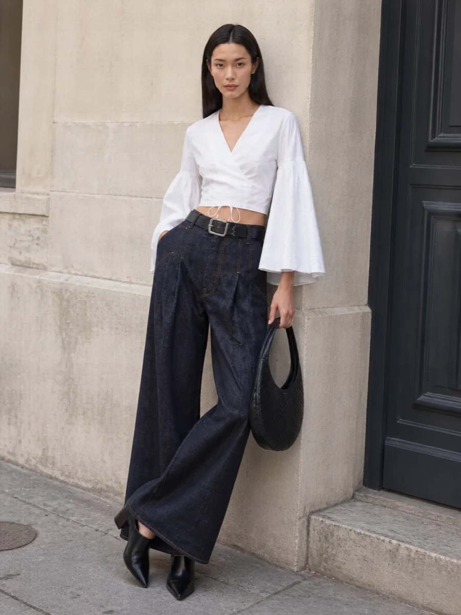 Dark indigo, ultra-wide Jacquemus denim is cinched with a sharp black belt and paired with the crisp white cropped wrap blouse, letting the dramatic bell sleeves and high waist carve a clean, architectural line. Black sculptural-heel mules add height and polish, while the sleek black Coperni shoulder bag quietly echoes the modern, minimalist geometry of the look for SS city evenings or gallery nights.
