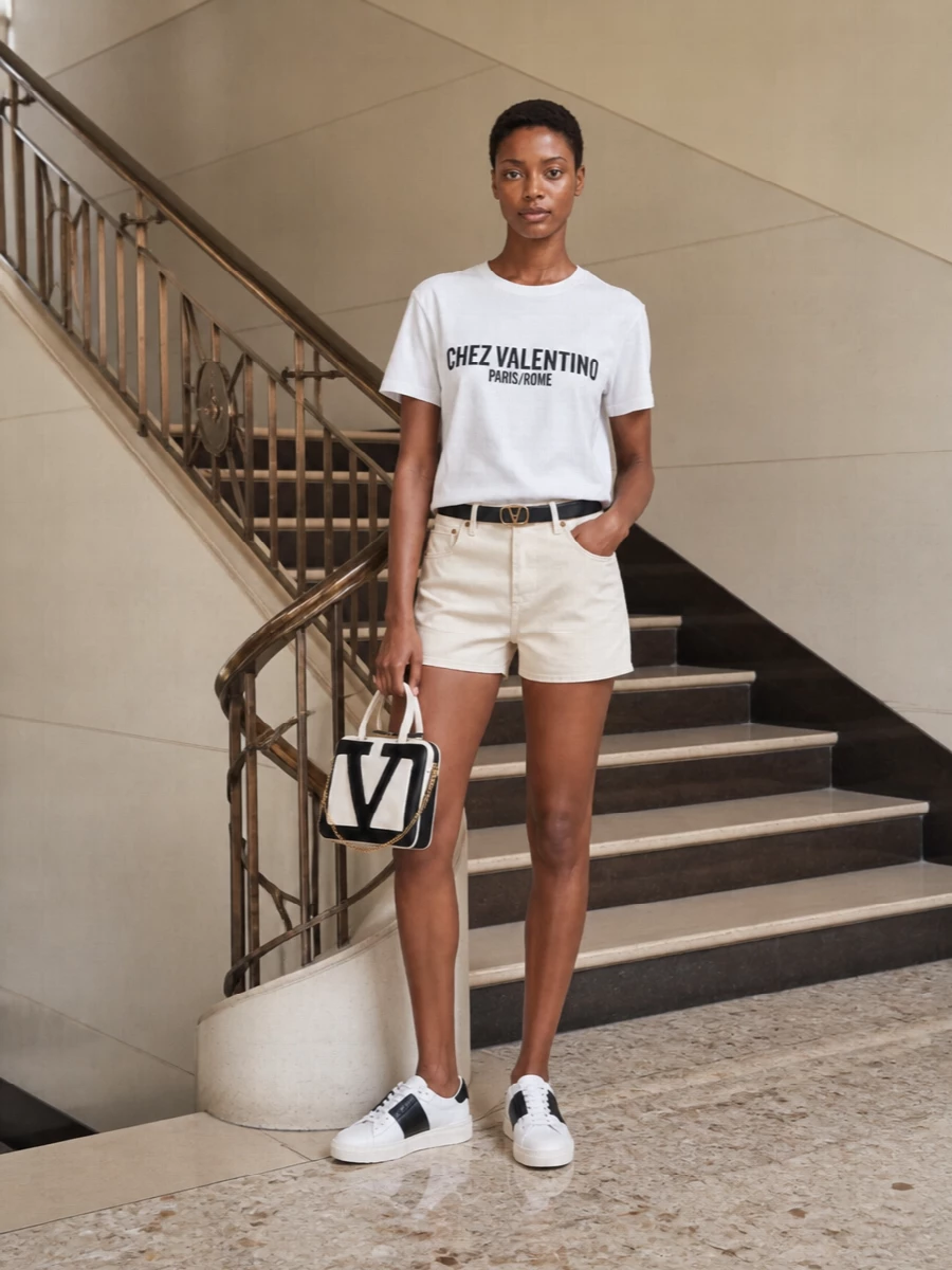 Graphic monochrome keeps this Valentino tee sharp, paired with cream tailored mini shorts for a clean, leggy proportion. White and black sneakers echo the chest logo, while a structured two tone tote and a slim VLogo belt add just enough Roman polish for city days. It is relaxed, precise and unmistakably designer without feeling dressed up.