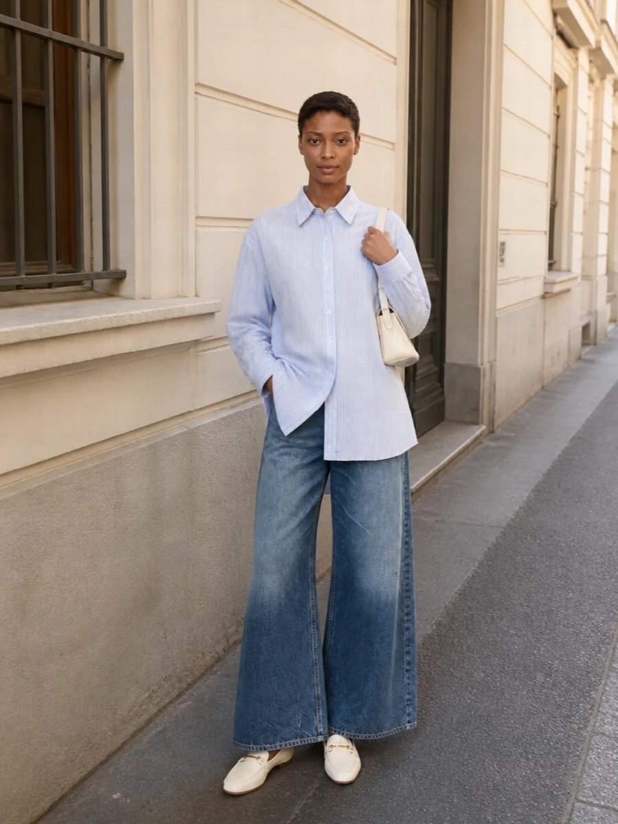 An oversized striped shirt and mid-rise wide-leg denim create an easy, elongated silhouette in layered shades of blue. Soft ivory loafers and a slim buttercream shoulder bag keep the palette airy and refined, subtly sharpening the relaxed proportions without breaking the quiet mood.