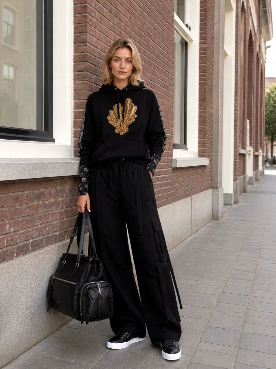 Dolce&Gabbana’s baroque hoodie is grounded with dark, wide-leg cargos so the metallic embroidery reads strong and modern rather than precious. Graphic low-top sneakers and a structured duffle keep the energy sporty and urban, while subtle logo sleeves add a final, tightly edited hit of branding. The result is a polished off-duty ensemble that feels dramatic yet easy for cold-city days.