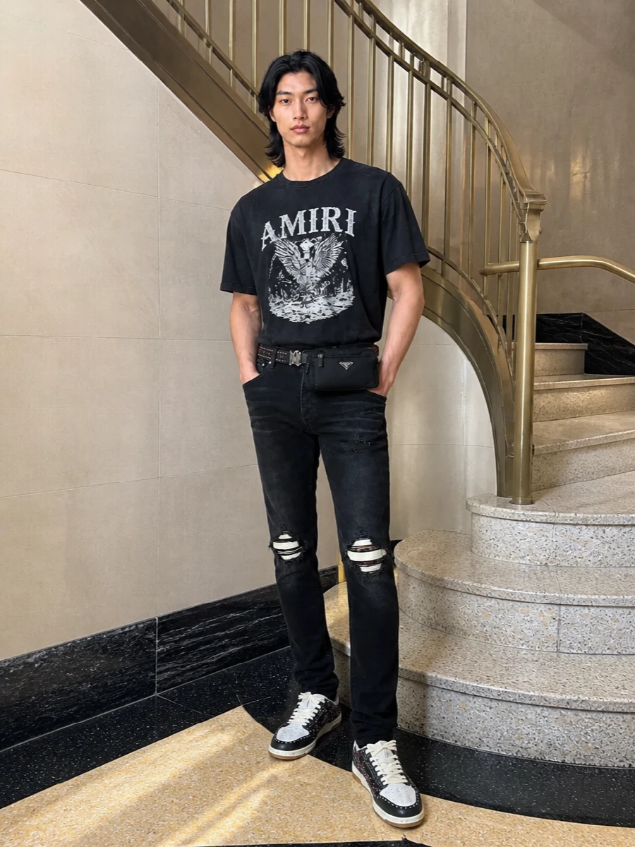 Jet-black ripped denim and a washed graphic tee set a tightly curated LA rock uniform, all in a deep monochrome base. Studded sneakers and the matching belt pull in flashes of ruby and metal, giving the outfit a deliberate, show-ready edge. The compact black belt bag keeps everything grounded and practical, so the attitude reads intentional rather than overdone.