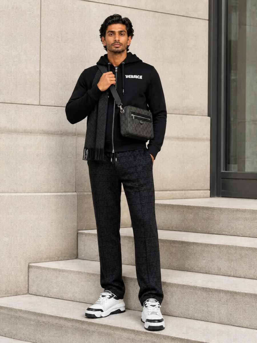 An all-black Versace hoodie and tonal-logo jogger form a clean, label-conscious base that reads polished, not tracksuit. Chunky off-white and black sneakers break the darkness just enough, while a charcoal monogram crossbody and GG scarf add quiet texture and city practicality. It is a streamlined streetwear uniform that feels intentionally luxurious from coffee runs to casual dinners.
