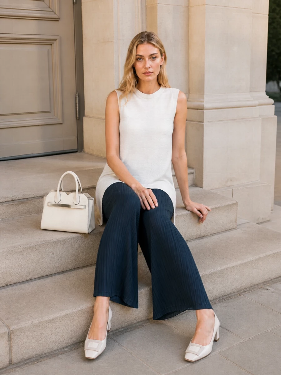 Soft white pleats skim over deep teal wide legs for a long, fluid line that feels calm yet considered. Ivory block-heel pumps and a sculpted Valextra bag keep the palette tonal and polished, while a whisper-light ivory scarf adds warmth without breaking the minimalist clarity.