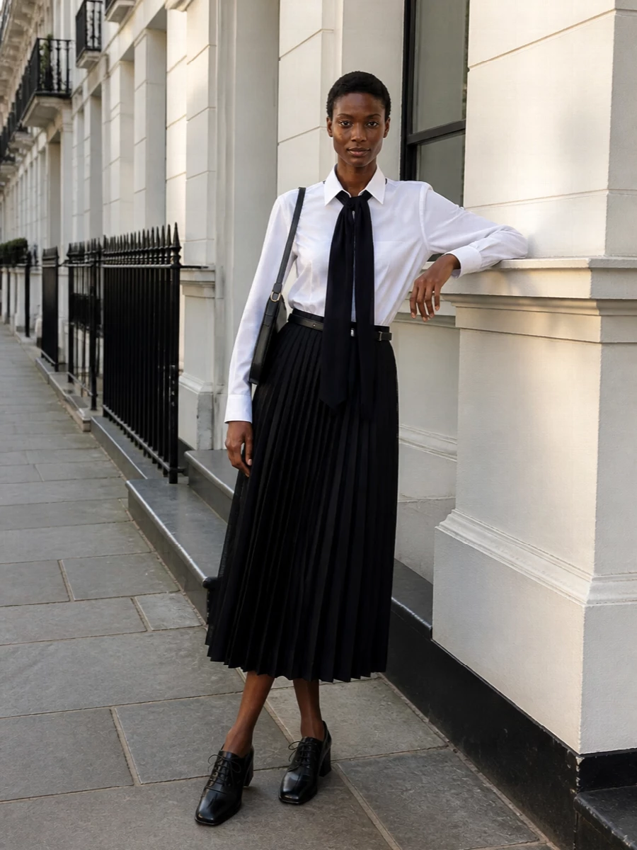 Crisp white and inky navy at the collar keep the focus on Sacai’s sculpted pleated skirt, creating a modern monochrome column that feels sharp but not severe. The square-toe lace-up heel and curved black shoulder bag reinforce the quiet, architectural mood, reading as considered from boardroom to late dinner. A slim black belt is the only punctuation at the waist, refining the silhouette without interrupting the skirt’s movement.