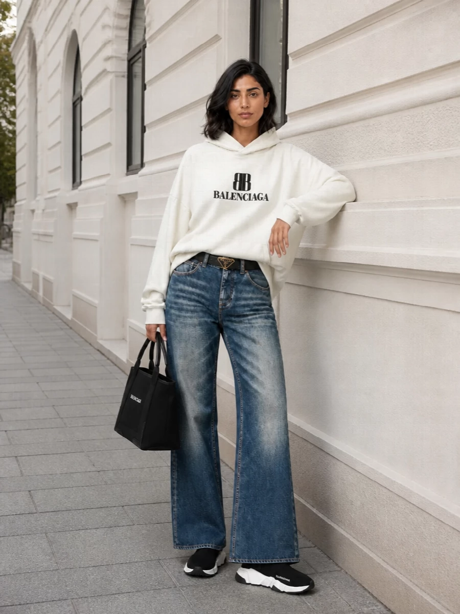 A faded wide-leg jean, oversized logo hoodie, and sock sneakers create a clean Balenciaga street uniform in off white, black, and worn indigo. The structured logo tote and slim black belt add just enough precision so the slouch reads intentional, not undone. It is relaxed, graphic, and quietly expensive without trying too hard.
