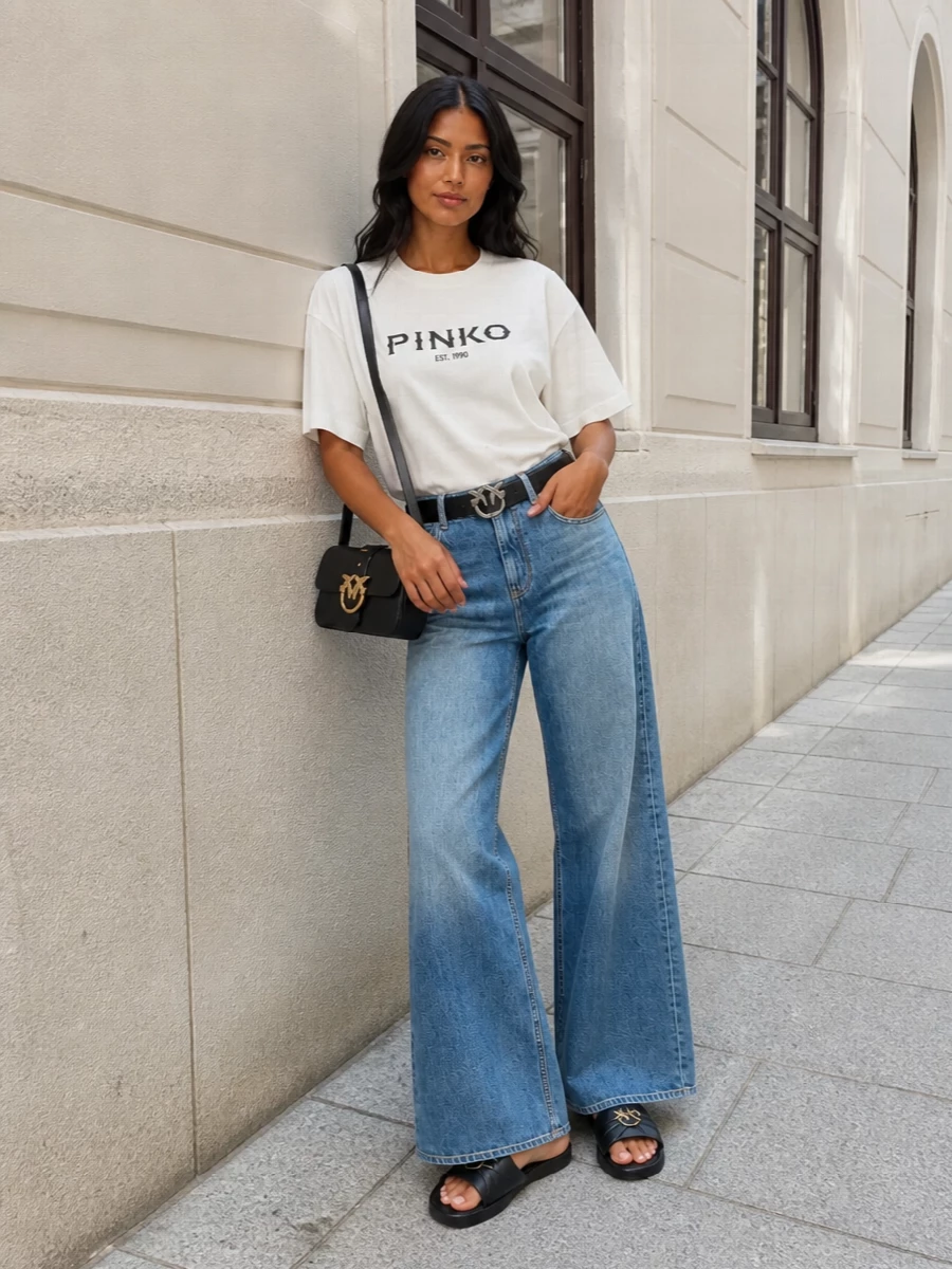 A graphic Pinko tee meets long, softened blue denim for a relaxed street take that still feels intentional. Black slides, a compact cross-body and a cinched waist keep the silhouette clean and anchored. The palette stays tight in white, blue and black, so the logo details read sharp rather than loud.