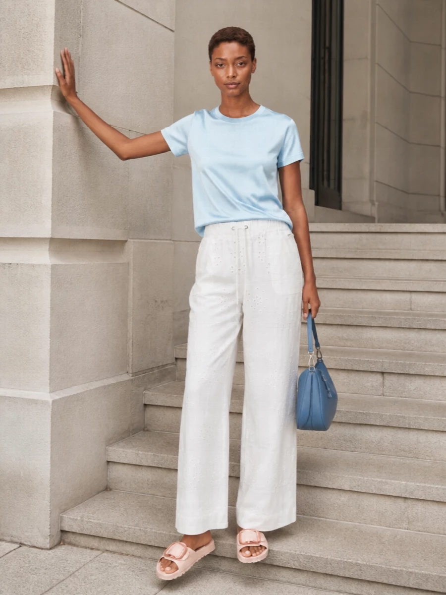 An ice-blue satin tee sits over white broderie wide-leg trousers for a mix of clean minimalism and soft, boho romance. Blush slides keep the mood relaxed and feminine, while the blue leather hobo quietly picks up the top’s cool tones. The result feels easy for warm days yet considered enough for terrace lunches or casual dinners.