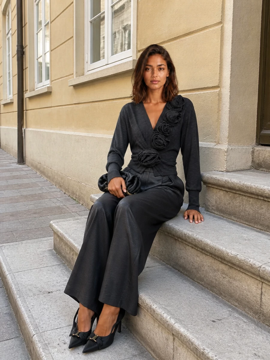Charcoal drape meets satin fluidity in a monochrome evening ensemble that feels romantic yet measured. The sculptural floral knit is grounded by long, high-waisted wide-leg trousers, creating an elongated, statuesque line. A sculpted bow slingback and rosette-like clutch quietly echo the floral story so the texture does the talking rather than obvious sparkle.