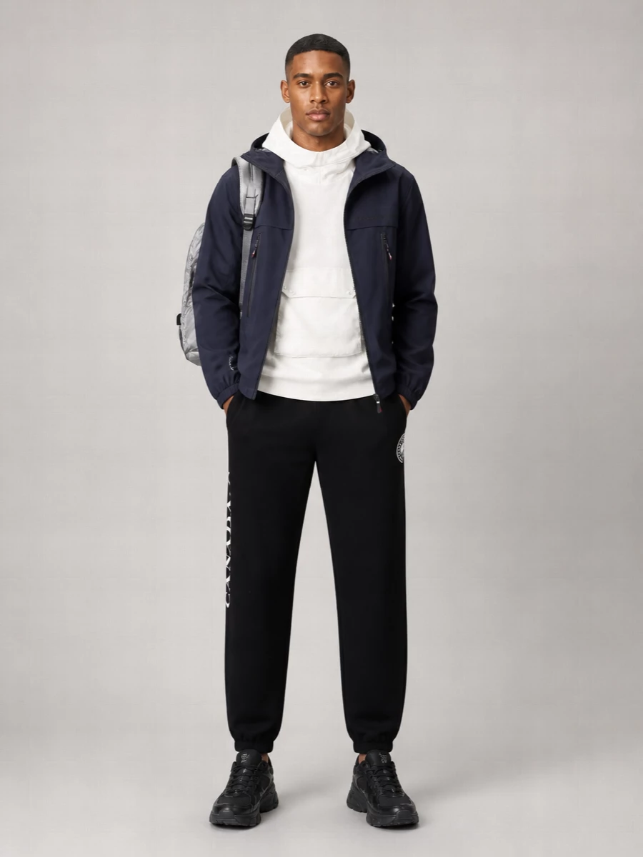 A navy technical shell, clean white hoodie and black track pant create a modern sport-uniform that moves easily from commute to weekend. All-black runners keep the lines sleek, while the reflective Stone Island pack adds a sharp, utilitarian contrast without breaking the minimalist palette.