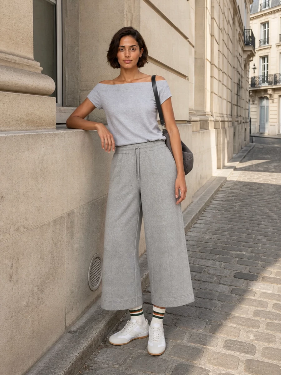 A tonal grey story keeps the mood calm and refined, with the off‑the‑shoulder knit and wide, cropped jersey trouser reading like a modern, relaxed set. Soft neutral runners and a charcoal crescent bag add just enough definition, keeping everything polished yet easy for city days or travel. The subtle striped sock detail gives a quiet nod to sport without disrupting the clean lines.