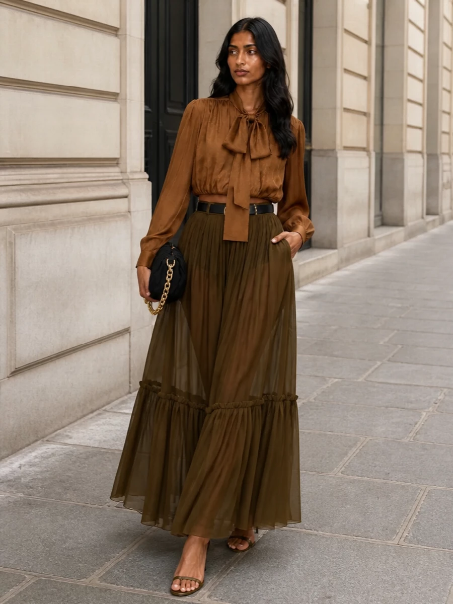 This look leans into a tonal, romantic SS evening story, with the sheer olive Saint Laurent maxi skirt paired to the caramel bow-neck blouse for a long, fluid line in warm neutrals. Olive-brown stilettos keep the palette seamless and legs sharp, while a black ruched pouch and slim black belt add just enough contrast and gold hardware to anchor the softness for polished, destination-ready nights out.