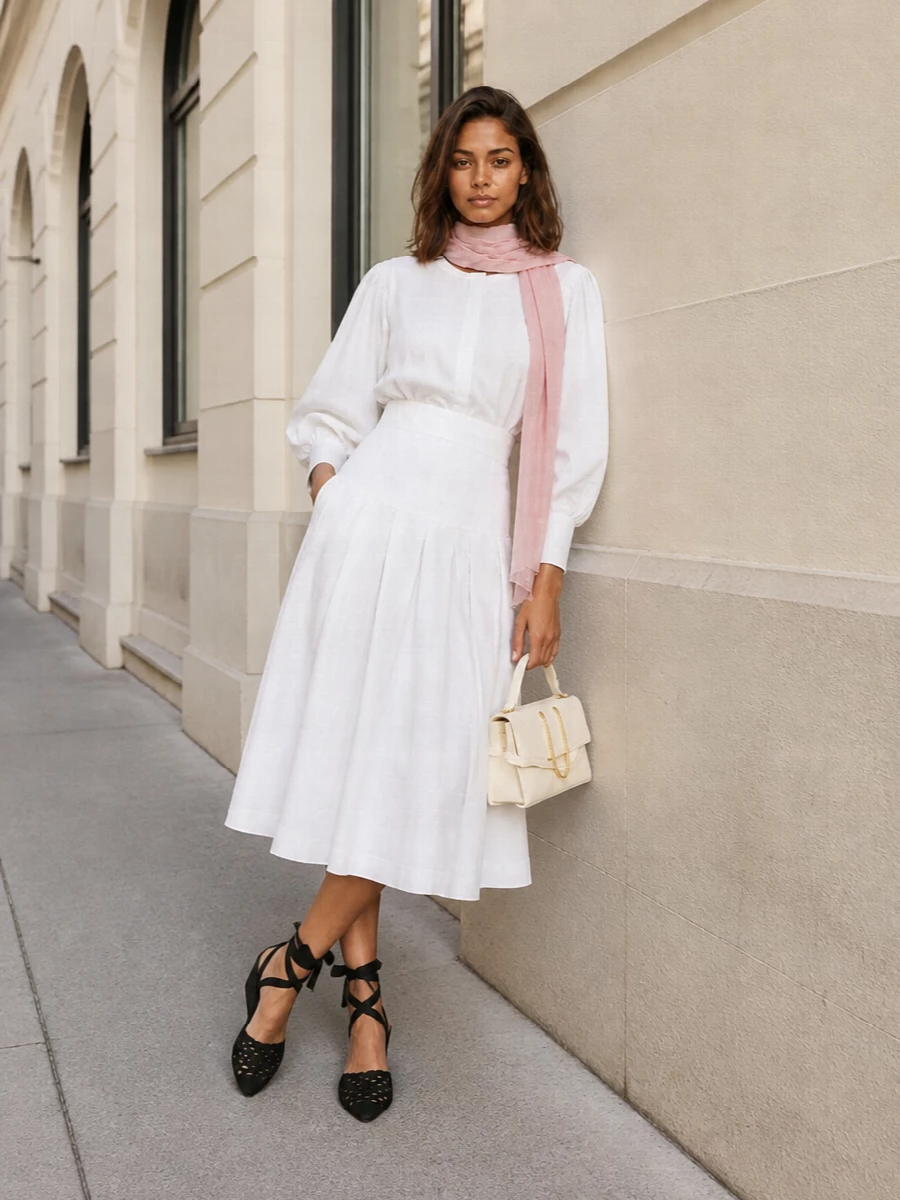 A sculptural white blouse meets a crisp pleated white midi for a monochrome silhouette that feels airy, architectural, and softly romantic. Black woven wedges and a cream top-handle bag add just enough contrast and texture to sharpen the mood while keeping it understated. A blush wrap at the neck or shoulders lends a gentle wash of color that stays firmly in the quiet luxury space.