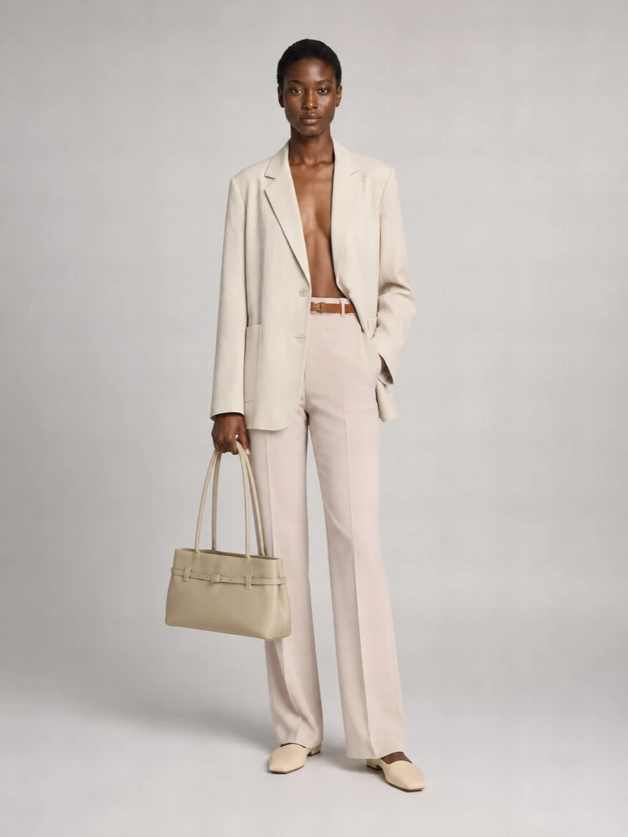 A soft-beige tailored column keeps the mood calm and precise, with the relaxed blazer and matching trouser reading like modern suiting rather than strict officewear. Buttercream flats and a stone top-handle bag stay within the same sun-washed neutral story, refining the silhouette without hard contrast. A slim camel suede belt quietly defines the waist so the proportions feel intentional, not slouchy.