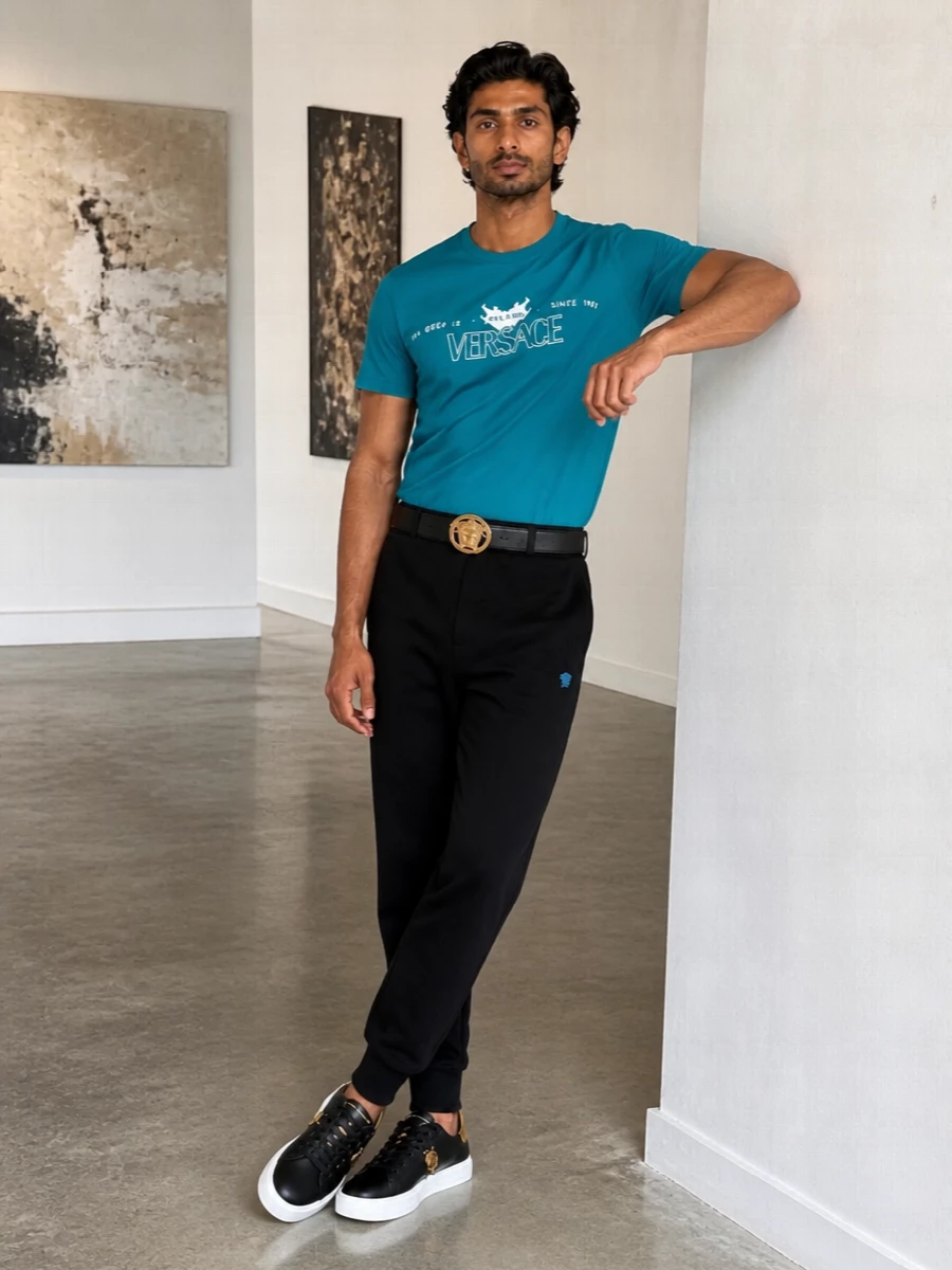 Teal and black do the talking here, with the logo tee grounded by clean cuffed joggers for a sharp, athletic streetwear feel. Gold hardware details run quietly through belt, sneakers and bag, giving the outfit a cohesive Versace edge without overwhelming the graphic color hit. It is the kind of off-duty uniform that reads confident and put together from airport lounge to late-night city laps.