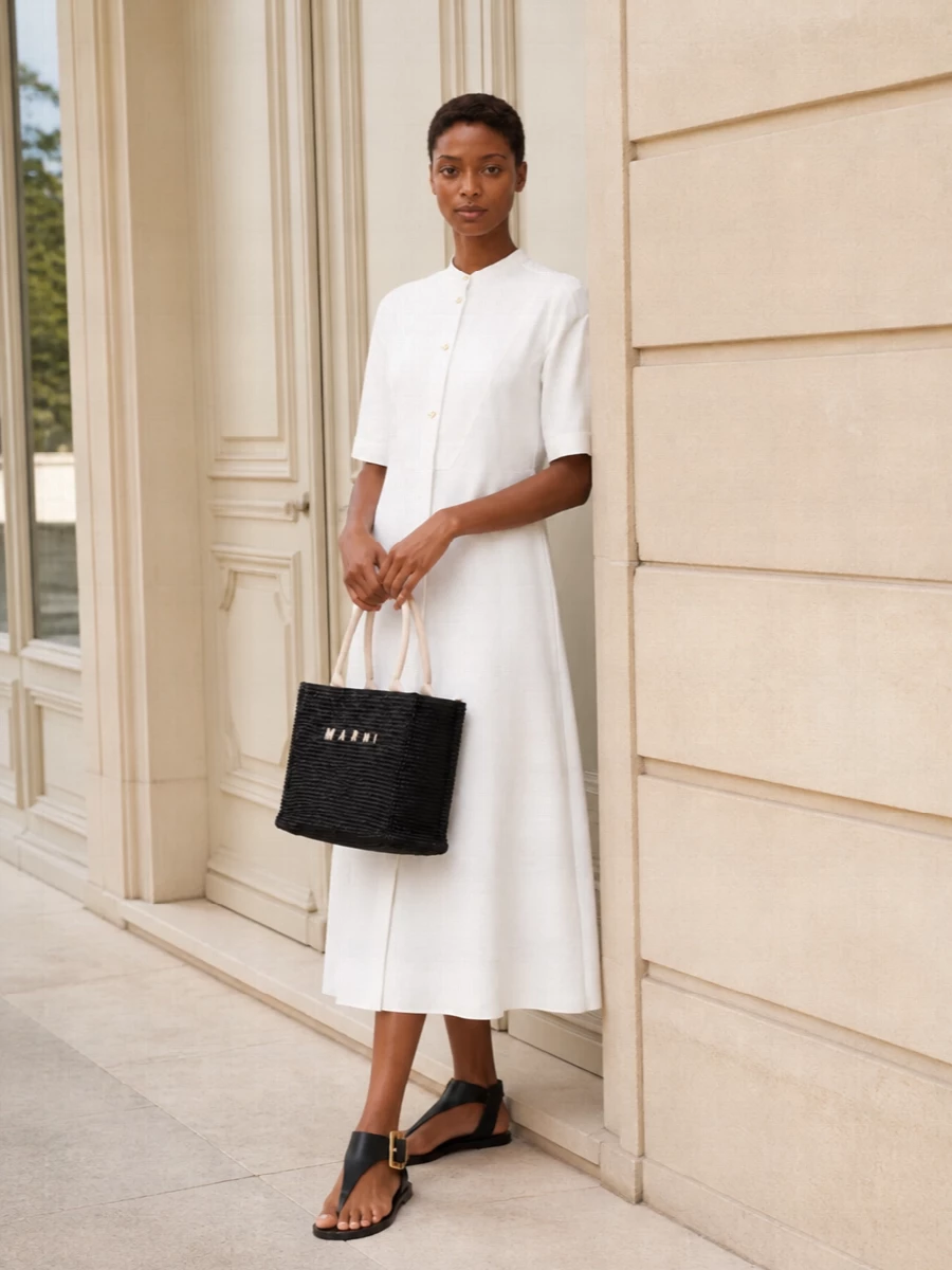 The Jil Sander shirt dress, grounded with black Balmain thong sandals, creates a clean summer column where the woven Marni tote becomes the tactile focal point. Soft white and black keep the palette graphic yet understated, perfect for city days or terrace dinners. Choosing the dress route over separates keeps the story minimal and lets the bag do the textural work without competing lines at the waist.
