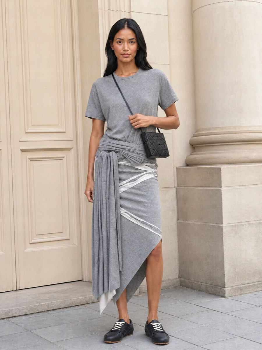 A sculptural grey tee and matching asymmetric skirt read like a single, wrapped column in soft jersey, all in a tight tonal palette. Chunky low-top sneakers keep the mood grounded and street, while the faceted cross-body bag adds a graphic, architectural hit that sharpens the softness. Wear it for city days when you want comfort to feel quietly intentional and a bit art-school cool.