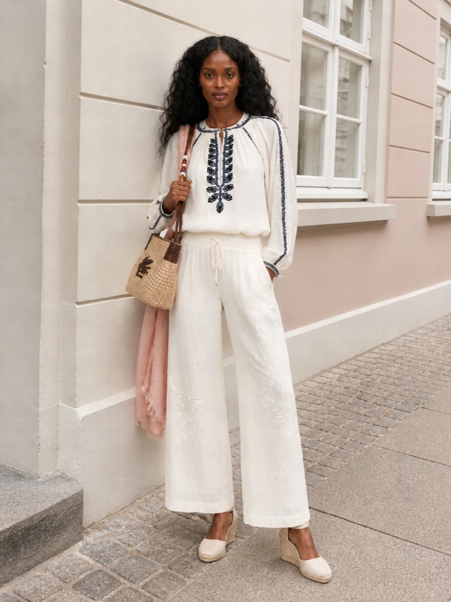 Soft ivory and navy embroidery meet fluid ivory trousers for a romantic, sun-ready outfit that feels relaxed but considered. The high waist and wide leg create a long, easy column, while the woven raffia shoulder bag and ivory wedges keep everything grounded in warm-weather boho charm. A blush scarf introduces just enough color for a quietly feminine finish.