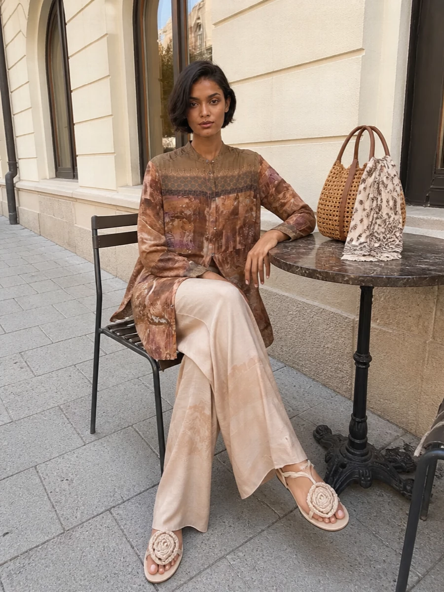 A long, tapestry-print shirt slips over ombré sand wide-legs for a soft, elongated column of warm neutrals. Cream lace-up sandals keep the mood relaxed yet refined, while a woven raffia tote and a chocolate‑toned scarf add texture and depth without disturbing the palette. The result is artistic and languid, perfect for warm days that move from gallery wandering to late dinners.