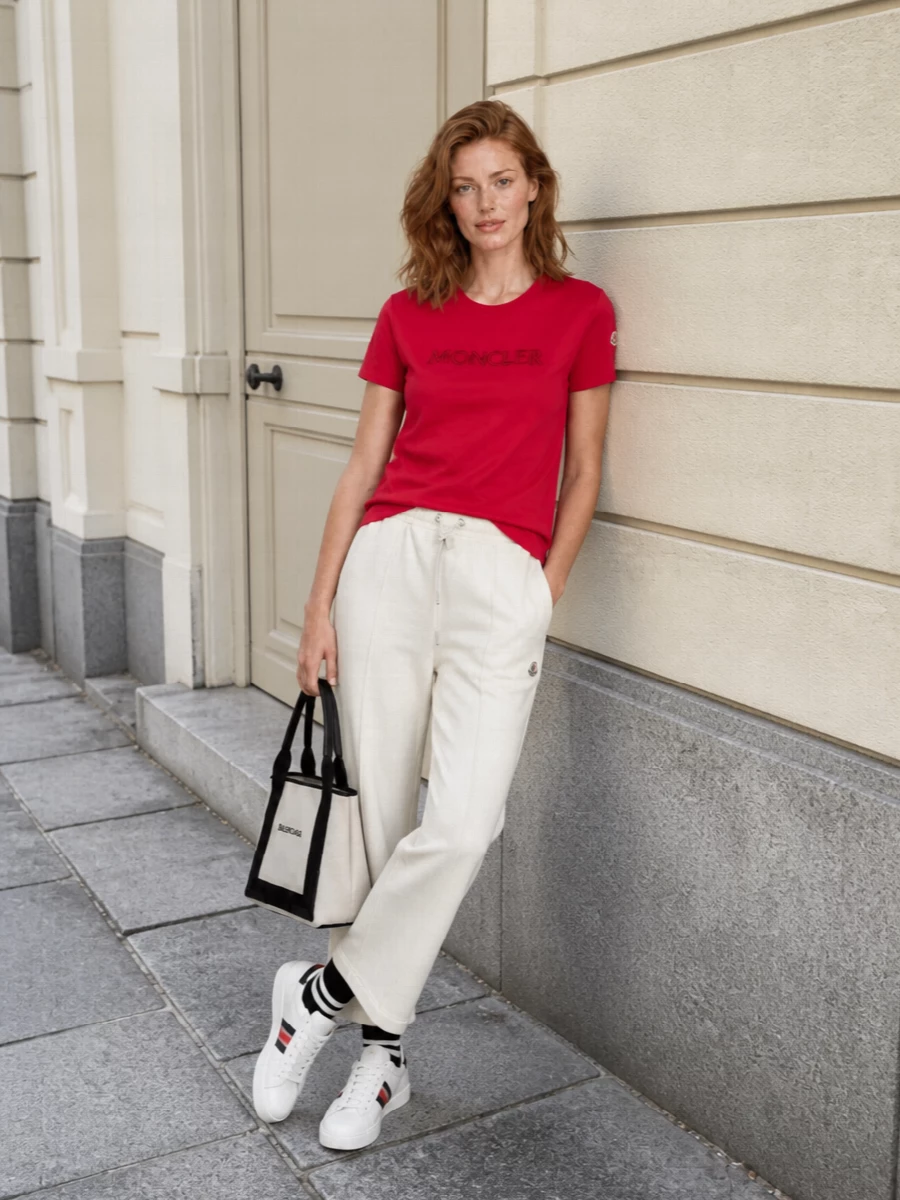 A vivid cherry Moncler tee is grounded with refined off‑white track pants for a clean, athletic take on off‑duty dressing. Tricolour court sneakers and sharp logo-accented socks keep the palette cohesive and sporty, while a structured Balenciaga mini tote adds just the right amount of architectural polish. The result is an elevated, SS-ready city look that moves easily from travel days to weekend errands.