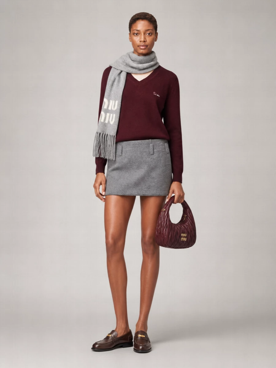 A deep burgundy V-neck meets a sharp grey mini for a uniform-adjacent take on quiet Miu Miu polish. Dark brown penny loafers ground the proportions, while the quilted burgundy bag picks up the knit’s richness for a concise, grown-up palette that still feels young and collegiate.