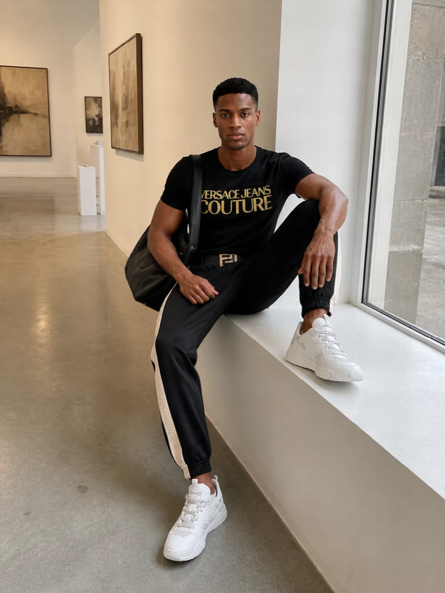 Head-to-toe Versace keeps this off-duty outfit tight, with the gold logo tee playing directly into the striped black joggers. Chunky white sneakers and the oversized leather shoulder bag bring a clean, athletic contrast, while the gold-buckled belt gives just enough structure to feel considered without losing ease.