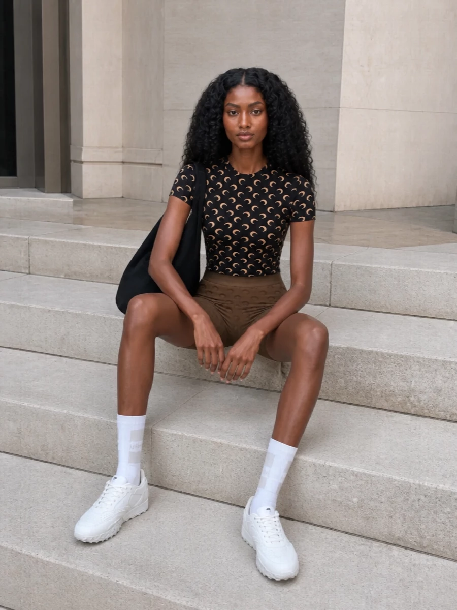 Marine Serre’s moon-print top and coordinated bike shorts read like a graphic second skin, all sharp curves and clean compression. White performance-style sneakers and a sculptural black tote keep it firmly in the street-sport world, practical enough for all-day wear but still very insider. The subtle hit of a white athletic sock finishes the palette with a precise, gym-adjacent nod rather than a fashion stunt.
