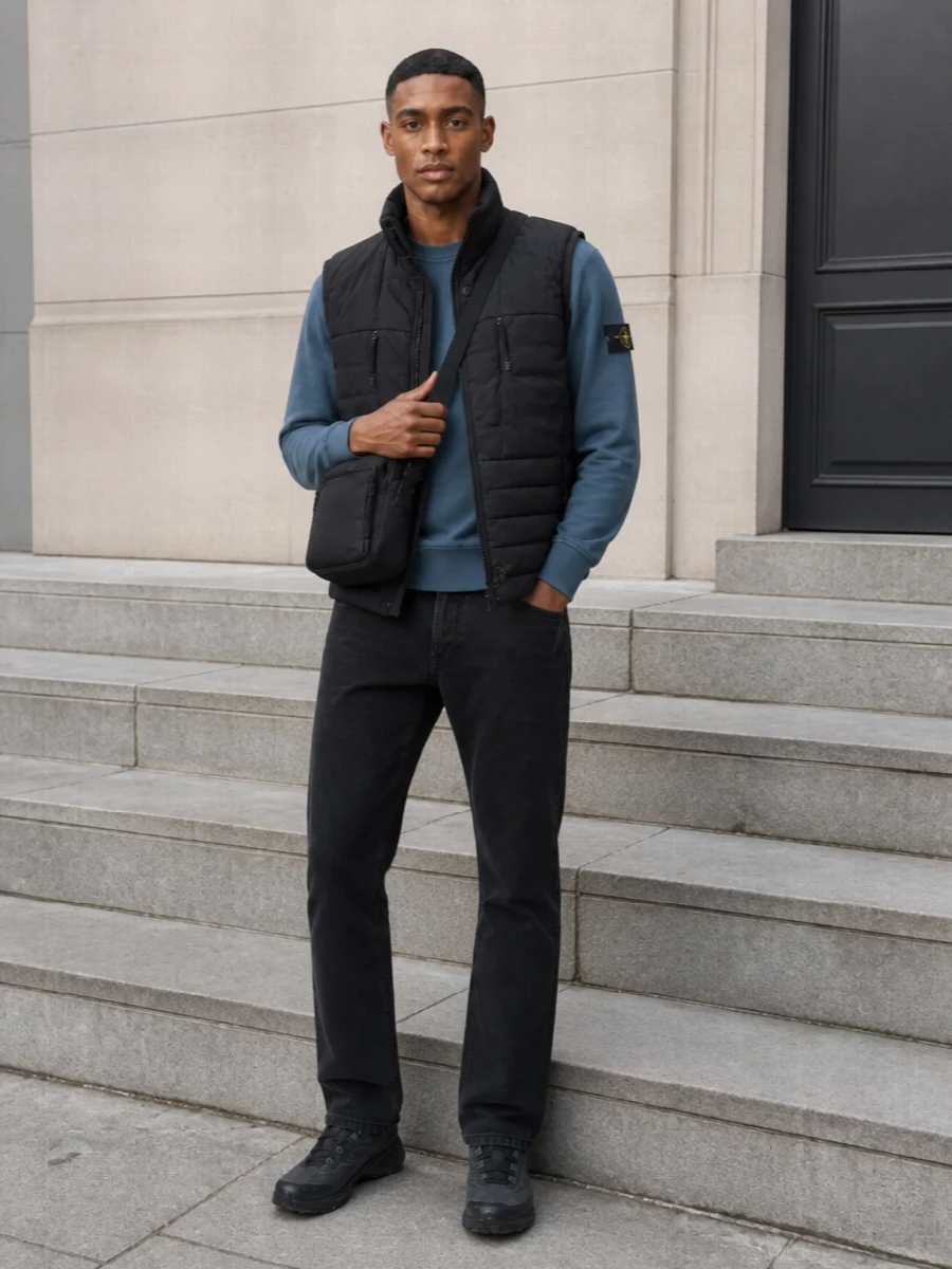 Stone Island’s padded gilet sits over a slate blue crew and washed black denim for a tight, technical street mix that feels considered but unfussy. All-black Salomon trail runners and a compact Stone Island crossbody keep the palette dark and functional, ready for cold city days and travel. The proportions stay clean and regular, so the volume of the vest is balanced by straight legs and low-profile accessories.