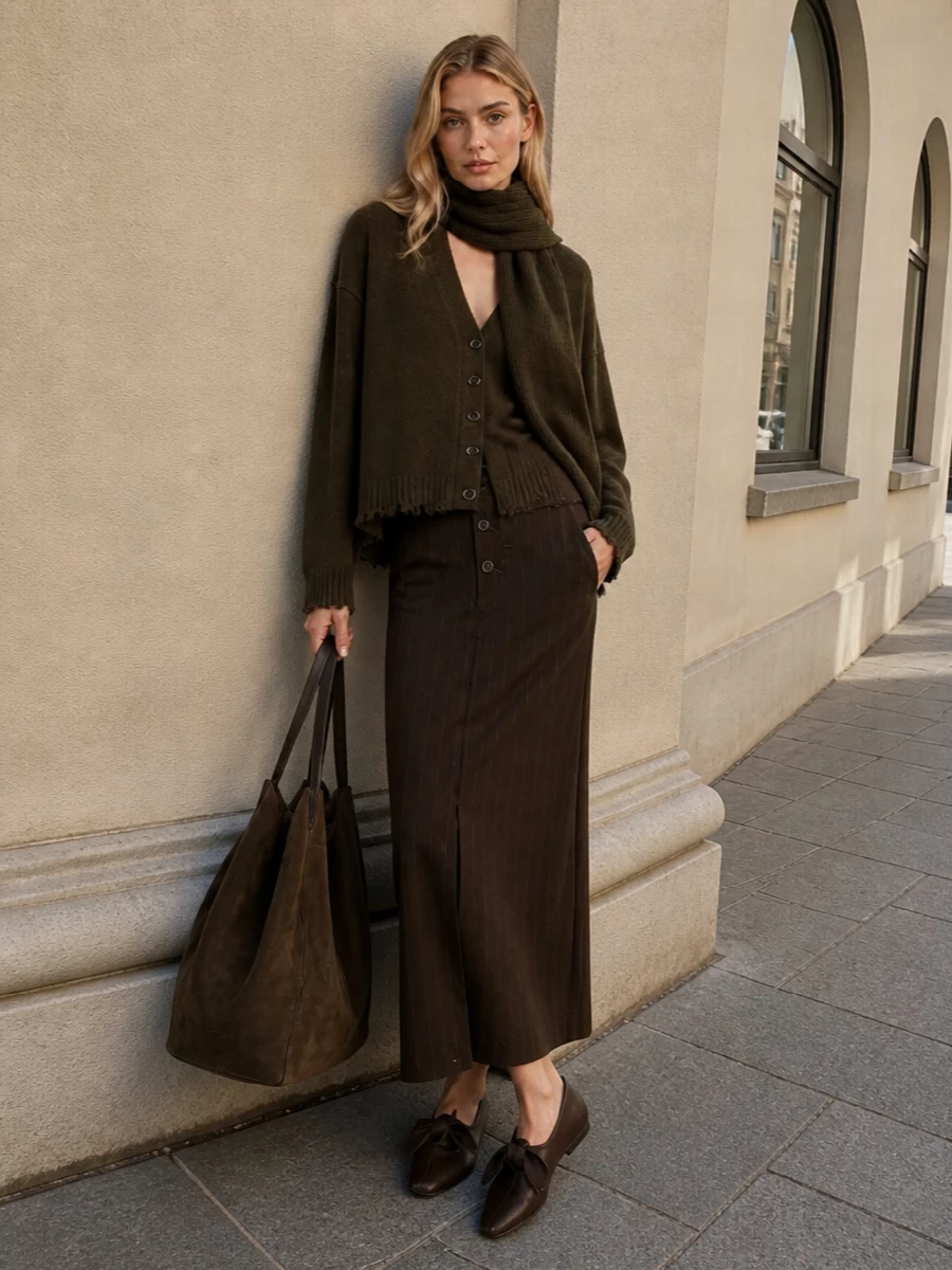 Deep olive and chocolate brown stack into a long, quiet column, with the pinstriped skirt sharpening the cardigan’s slouch into something quietly precise. Sculptural flats and a substantial suede tote keep the mood grounded and tactile, an earthy mix of tailoring and softness that reads thoughtful rather than fussy. The matching olive scarf is a final, subtle layer of texture that ties the tones together around the face.