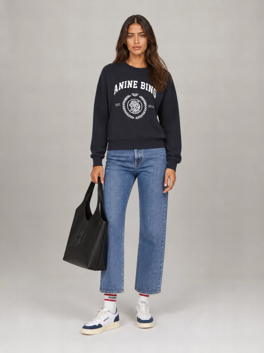 Anine Bing varsity energy stays sharp with high-rise straight blue denim that keeps the proportions long and clean. Navy and off-white sneakers and a sculpted black tote lean into a retro campus mood while still reading grown up. Striped sport socks add a small hit of color play that ties the whole palette together without competing with the crest graphic.