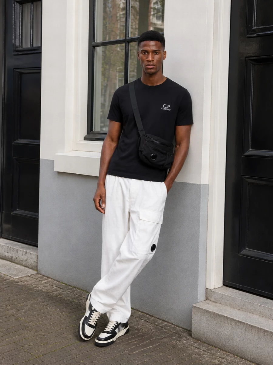 A black C.P. Company tee over loose white technical cargos creates a crisp, monochrome streetwear uniform with an easy, athletic slouch. The Represent sneakers pick up the same black and off‑white contrast, while the compact C.P. Company crossbody and discreet Ami socks keep everything feeling considered and urban without breaking the clean lines.