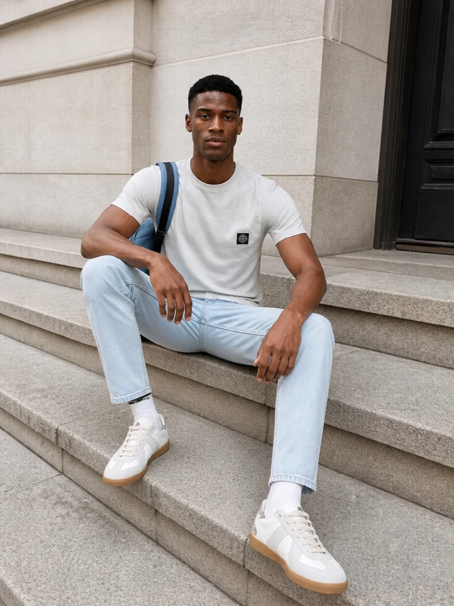 A pale grey Stone Island tee sits over icy straight-leg denim for a clean, tonal base that feels sharp but unfussy. Margiela’s soft white and grey sneakers keep everything light underfoot, while the slate blue technical backpack adds just enough structure and function. A subtle striped sock detail nods to retro sport, tying the streetwear mood together without crowding the palette.
